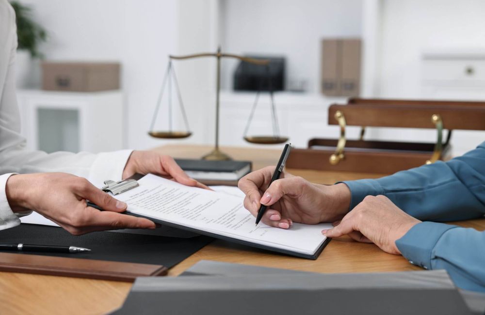 Two individuals are seated at a desk in a legal office, with one person handing over a document on a clipboard while the other signs it. A set of scales of justice is visible in the background, symbolizing the legal process. This visual represents the formal procedure of appointing personal representatives to manage the estate of someone who dies without a will in Virginia.