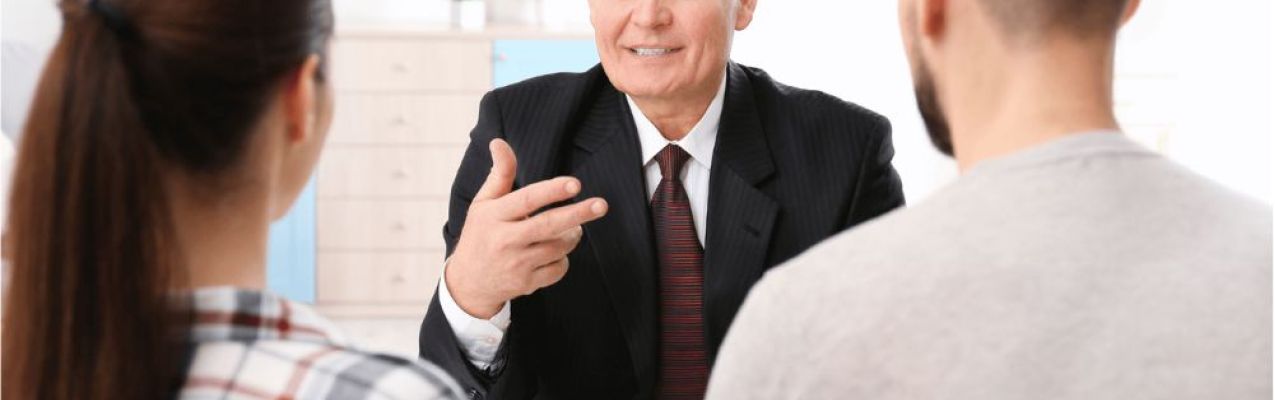 A man in a suit is seated at a table, gesturing and speaking to a couple, who are attentively listening. The room is bright and includes decorative plants.