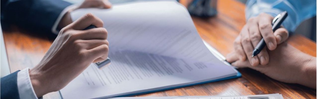 A hand holds a pen, poised to sign a document on a wooden desk. Nearby, a calculator and printed reports are visible. Bright daylight filters through a window, adding warmth.
