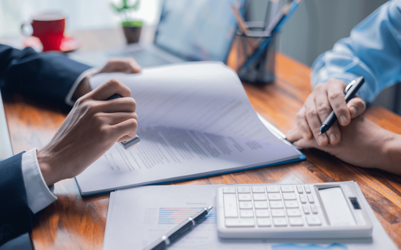 A hand holds a pen, poised to sign a document on a wooden desk. Nearby, a calculator and printed reports are visible. Bright daylight filters through a window, adding warmth.