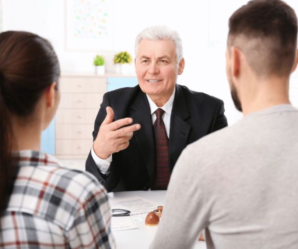 A man in a suit is seated at a table, gesturing and speaking to a couple, who are attentively listening. The room is bright and includes decorative plants.