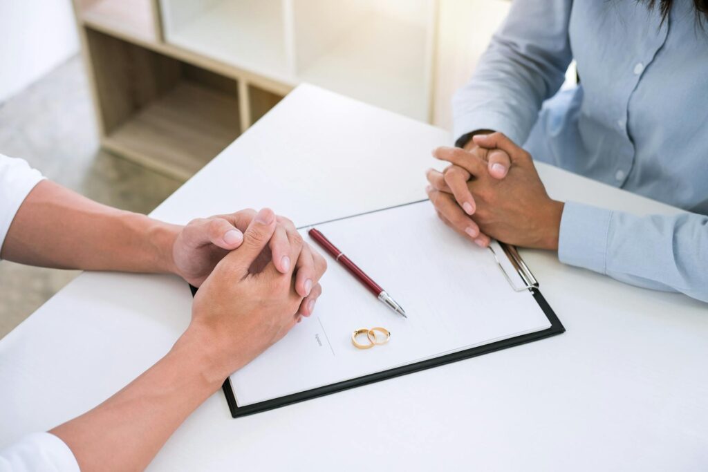 What Factors Determine Whether Your Ex-Spouse Gets Your Inheritance - Virginia Beach Law Group Two pairs of hands rest on a table, one set clasped, while the other holds a pen over a document. A pair of wedding rings is placed on the table, suggesting a discussion about marriage or divorce.